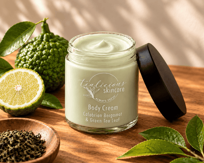 Open jar of pale green Calabrian Bergamot & Green Tea Leaf Body Cream beside fresh bergamot, tea leaves and a black lid.