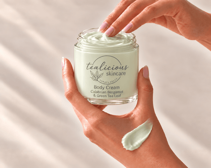 Hands holding an open jar of Calabrian Bergamot & Green Tea Leaf Body Cream, with a fingertip lifting cream and a pale green swatch on the hand.