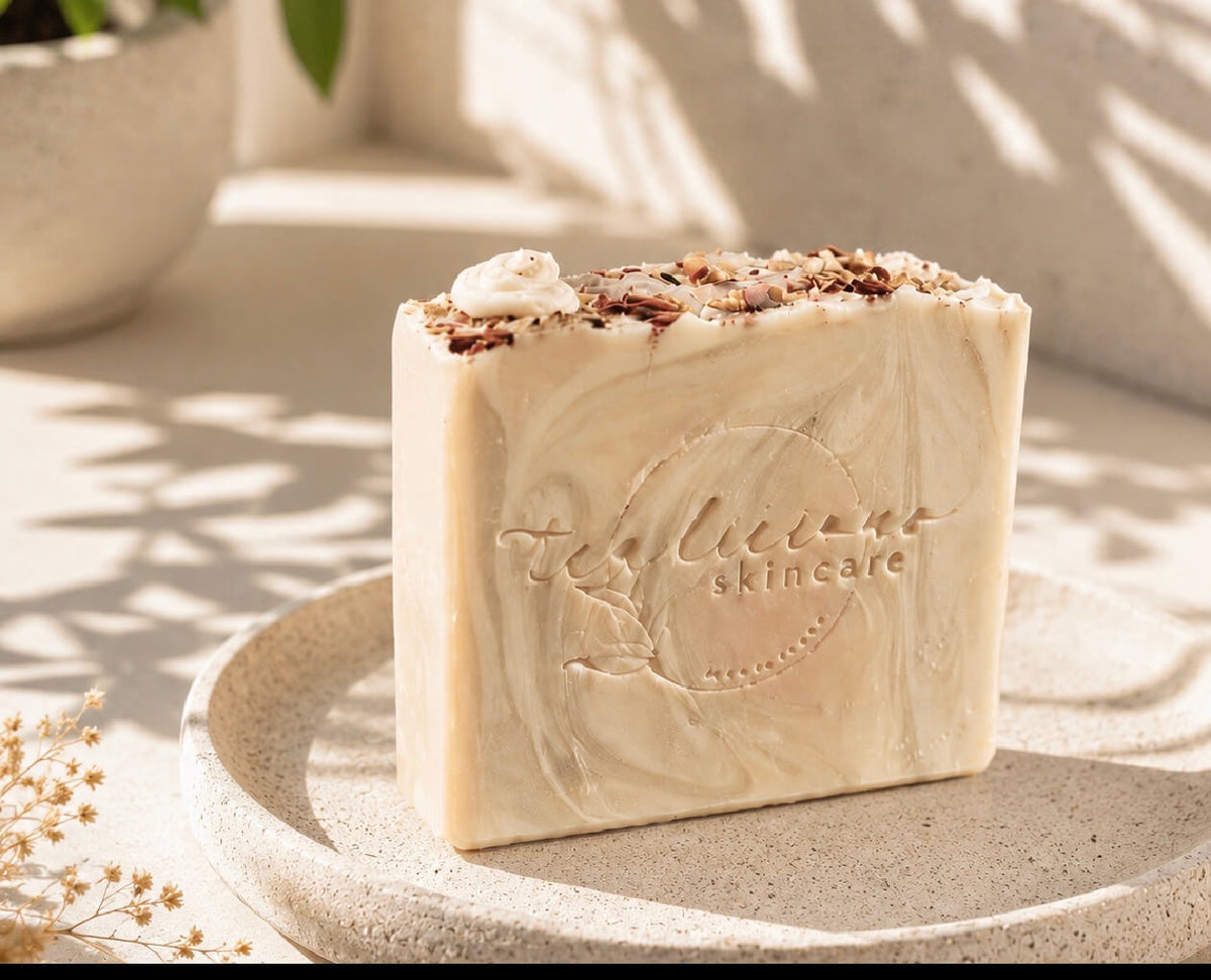 Cream-colored Tea-Licious soap bar with an embossed logo and botanical topping, displayed on a stone tray in soft sunlight.