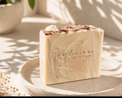 Cream-colored Tea-Licious soap bar with an embossed logo and botanical topping, displayed on a stone tray in soft sunlight.