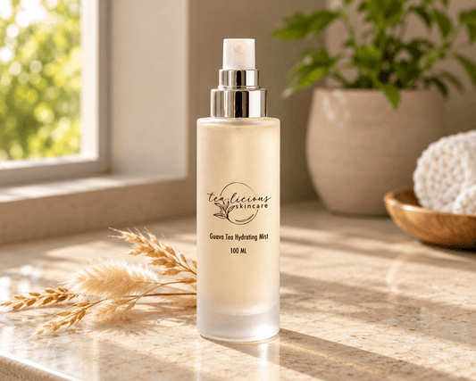 Tea-licious Guava Tea Hydrating Mist in a frosted glass spray bottle on a sunlit stone vanity beside dried grasses and a rolled cream towel.