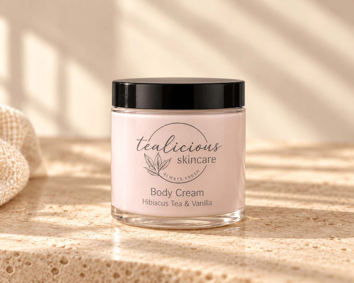 Tea-licious Hibiscus Tea & Vanilla Body Cream in a glass jar with a black lid, on neutral stone beside cream linen in soft natural light.