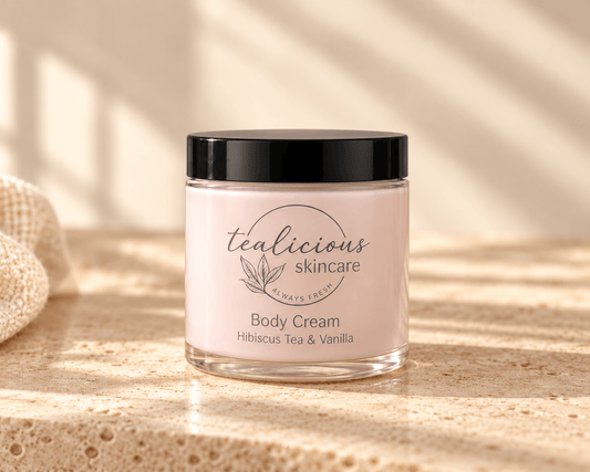 Tea-licious Hibiscus Tea & Vanilla Body Cream in a glass jar with a black lid, on neutral stone beside cream linen in soft natural light.