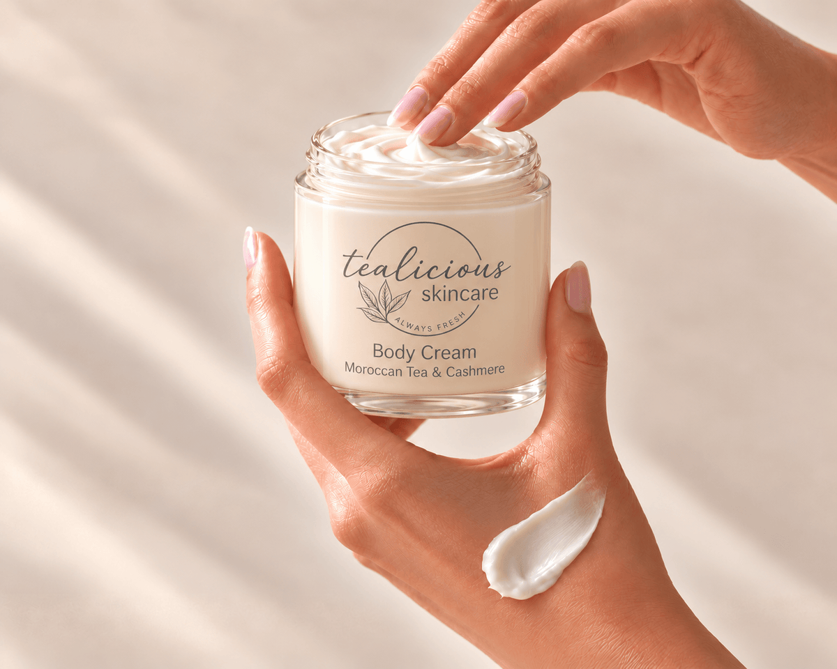 A person holds a clear glass jar of Tealicious Skincare Body Cream, with cream visible inside and a dollop on the palm, against a warm beige background with soft shadows.