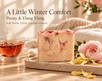 Tea-licious Peony & Ylang Ylang soap on a stone tray beside warm tea, soft towels and flowers. Text reads “A Little Winter Comfort. Soft florals. Clean, graceful comfort.”