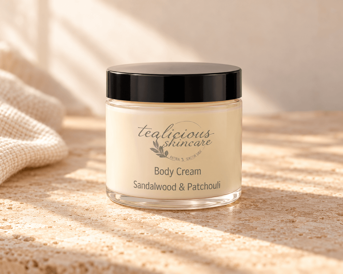 Tea-licious Sandalwood & Patchouli Body Cream in a glass jar with a black lid, on neutral stone beside cream linen in soft natural light.