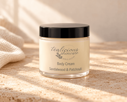 Tea-licious Sandalwood & Patchouli Body Cream in a glass jar with a black lid, on neutral stone beside cream linen in soft natural light.