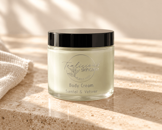 Tea-licious Santal & Vetiver Body Cream in a glass jar with a black lid and silver lettering, on neutral stone in soft natural light.