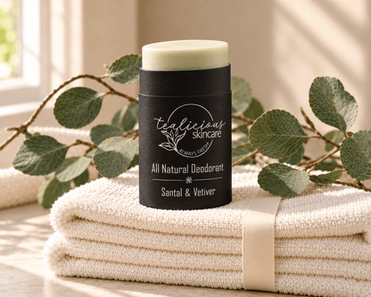Tea-licious Santal & Vetiver deodorant in a black oval container with silver lettering, on ivory towels tied with ribbon beside eucalyptus.