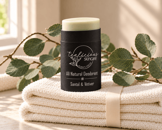 Tea-licious Santal & Vetiver deodorant in a black oval container with silver lettering, on ivory towels tied with ribbon beside eucalyptus.
