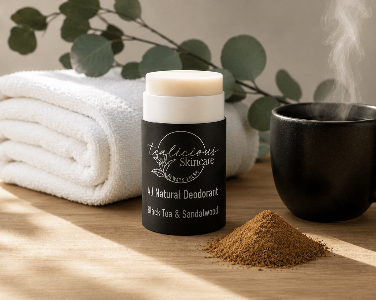 Black Tea & Sandalwood Deodorant stick with 'Tea-licious Skincare' branding on a wooden surface with a towel, mug, and leaves in the background.