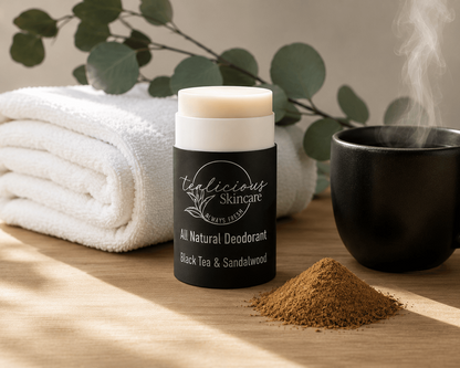 Black Tea & Sandalwood Deodorant stick with 'Tea-licious Skincare' branding on a wooden surface with a towel, mug, and leaves in the background.