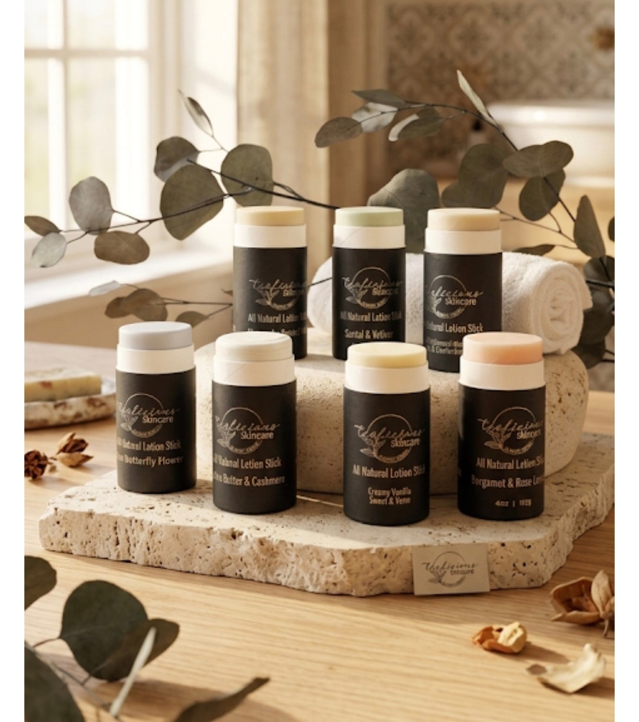  A collection of seven Tea-licious Skincare all-natural lotion sticks in various scents, including Blue Butterfly Flower and Santal & Vetiver, arranged on stone pedestals with eucalyptus leaves.
