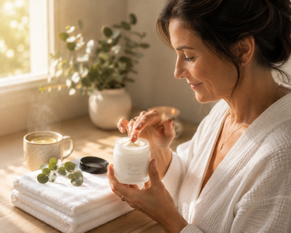 Woman in a white robe applying tea-licious skincare cream to her hand in a cozy room with plants and a cup.