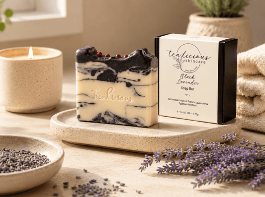 Tealicious Skincare Black Lavender Soap Bar with a cream and black marble pattern and small red berry accents, displayed next to its black and cream packaging box on a stone tray with lavender and a lit candle in a cozy setting.