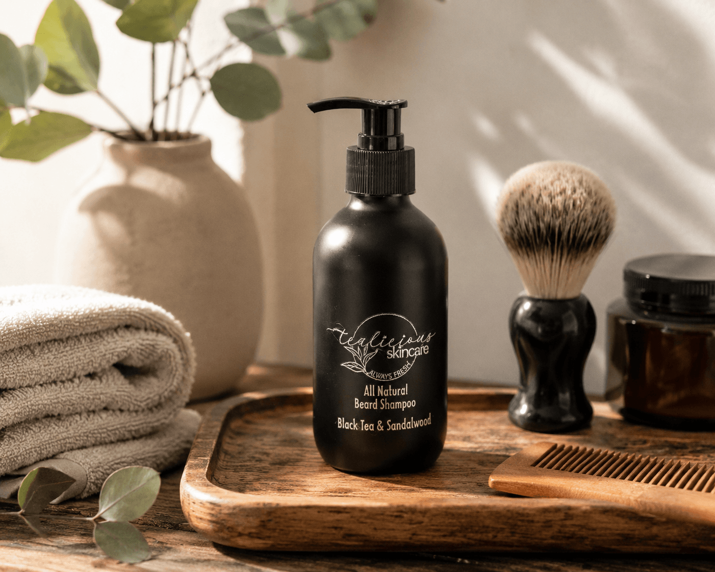 Tea-Licious Skincare Black Tea & Sandalwood Beard Shampoo displayed on a wooden tray with a shaving brush, wooden comb, folded towels, and eucalyptus accents in soft natural light.