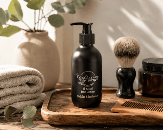 Tea-Licious Skincare Black Tea & Sandalwood Beard Shampoo displayed on a wooden tray with a shaving brush, wooden comb, folded towels, and eucalyptus accents in soft natural light.