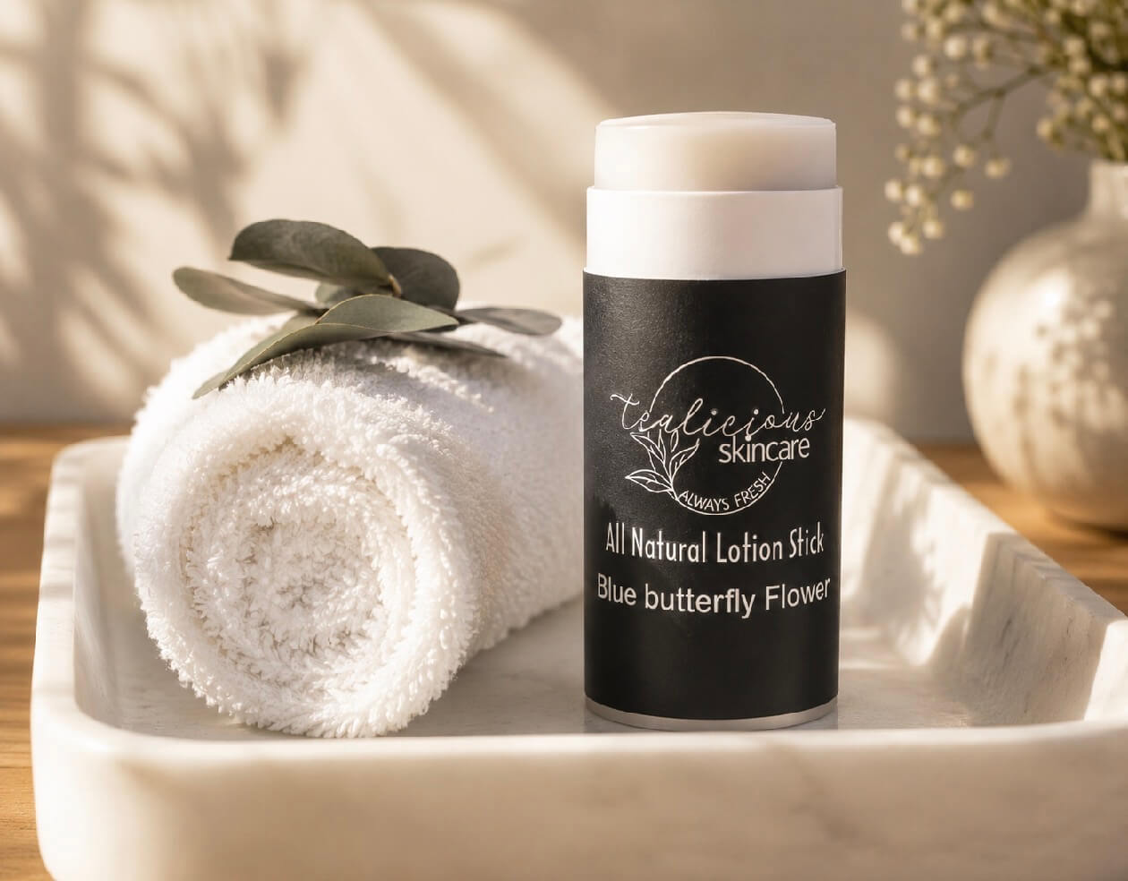 Tea-licious Skincare Blue Butterfly Flower lotion stick on a white marble tray next to a rolled white towel and a green leaf in a bright spa setting.