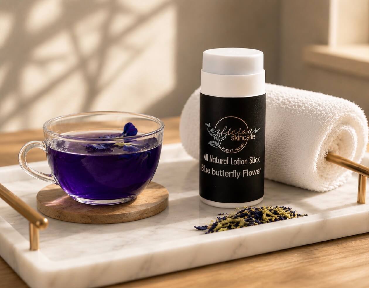 Tea-licious Skincare Blue Butterfly Flower lotion stick on a marble tray next to a cup of purple herbal tea and a rolled white towel.