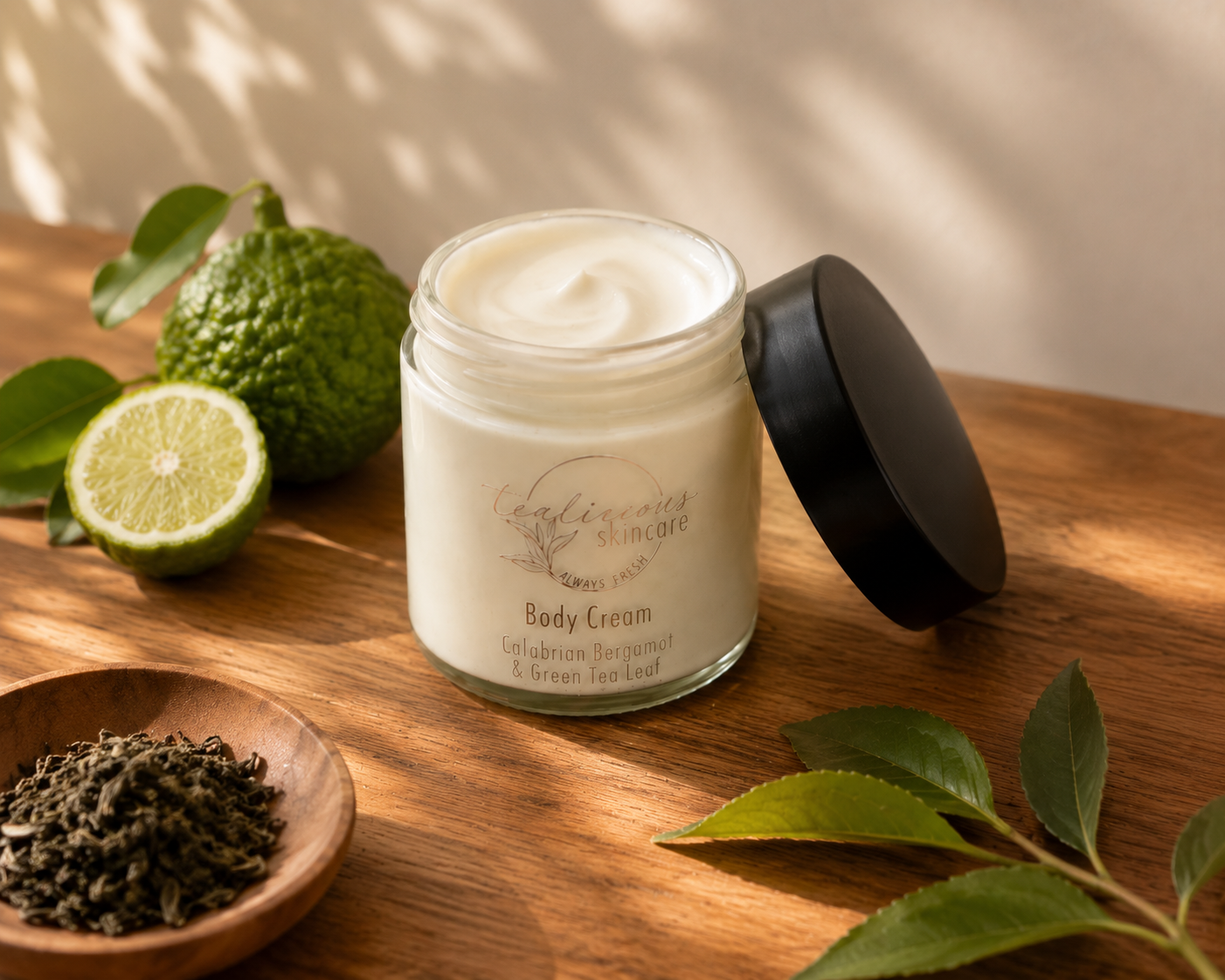An open glass jar of Tea-licious Skincare Calabrian Bergamot & Green Tea Leaf body cream sits on a wooden surface. The scene is captured in soft morning light with realistic soft-edged shadows, featuring a wooden bowl of dried tea leaves and fresh bergamot fruit in the background.