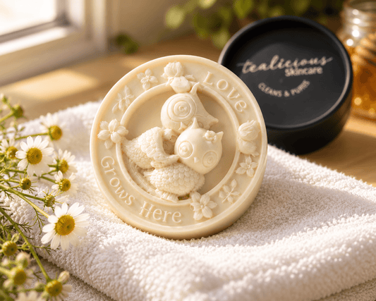 Tea-licious Chamomile & Honey owl massage bar on a white towel beside its black container, chamomile flowers and a honey jar.