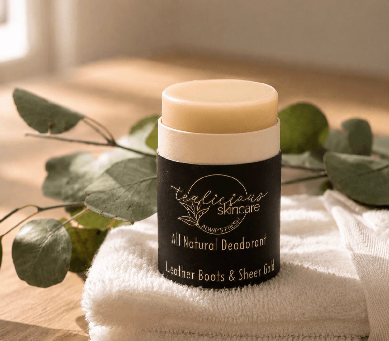 Tea-licious Skincare Leather Boots & Sheer Gold baking soda free deodorant styled with eucalyptus and a towel in warm morning light.