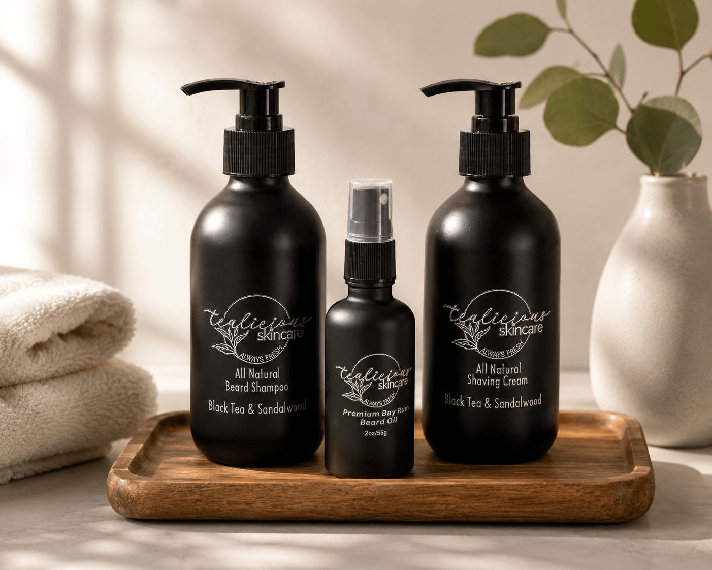 Tea-Licious Skincare men’s grooming collection featuring Black Tea & Sandalwood Beard Shampoo, Bay Rum Beard Oil, and Black Tea & Sandalwood Shaving Cream displayed on a wooden tray with folded towels and eucalyptus in soft natural light.
