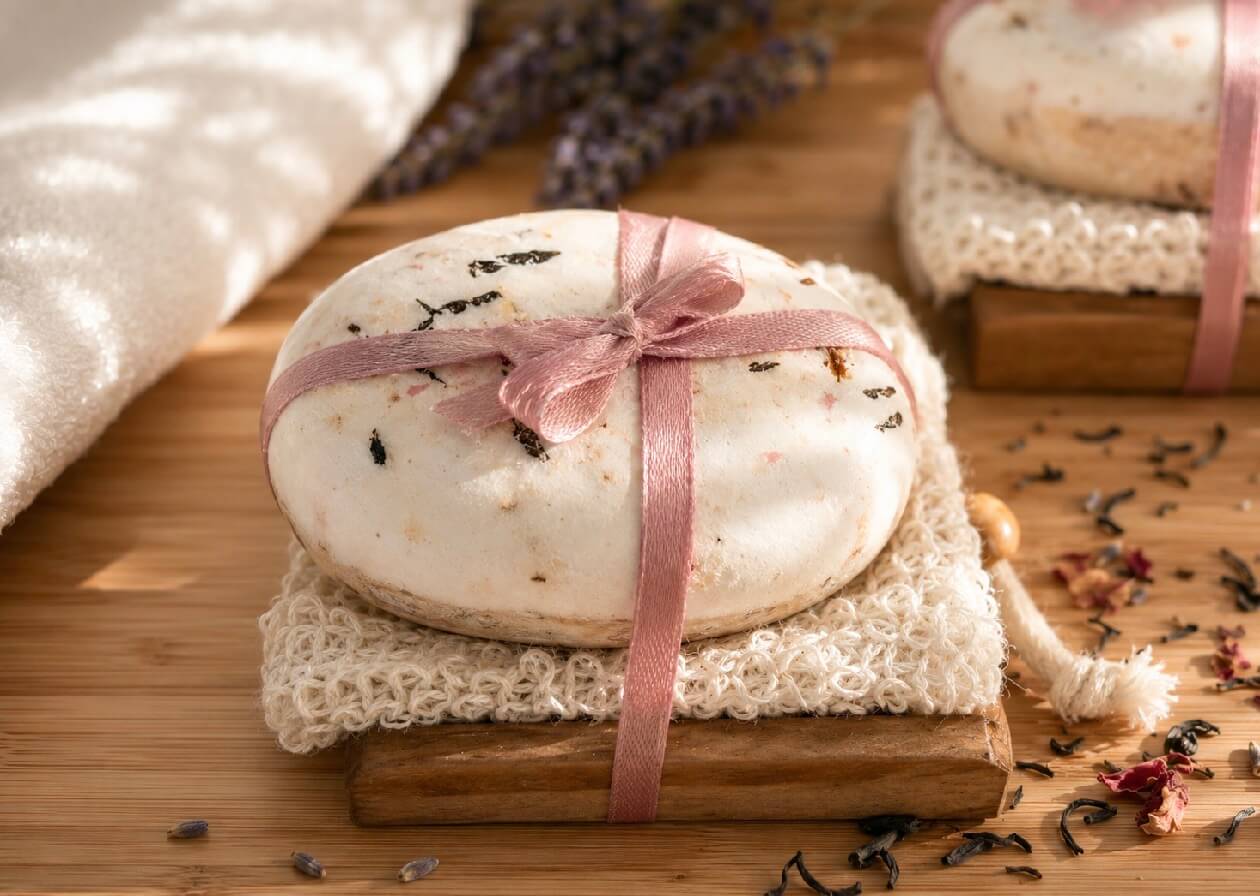 Tea-licious rose petal tea soap wrapped with pink ribbon on natural soap saver surrounded by tea leaves and botanicals.