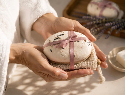 Hands holding Tea-licious rose petal tea soap wrapped with ribbon on a natural soap saver in warm spa lighting.