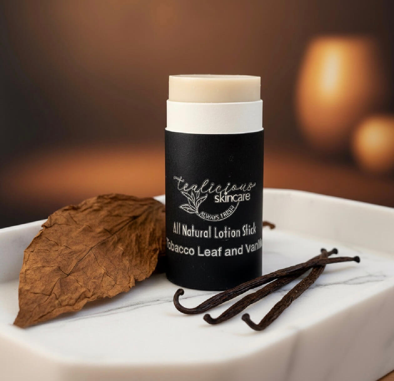 A Tea-licious Skincare Tobacco Leaf and Vanilla lotion stick on a marble tray with a dried tobacco leaf and vanilla beans.