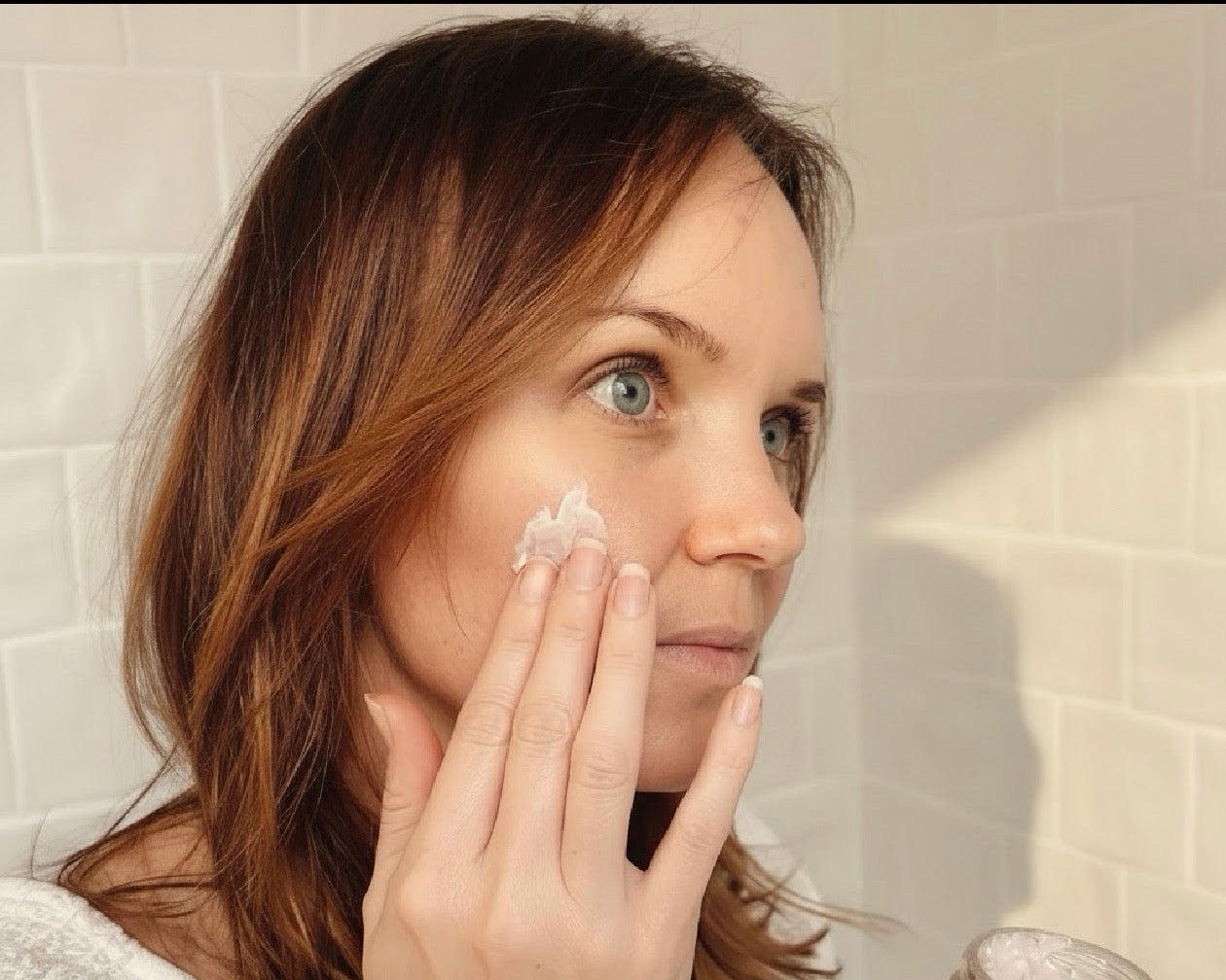 Woman applying White Guava Tea Moisture Infusion cream in soft natural bathroom morning light.
