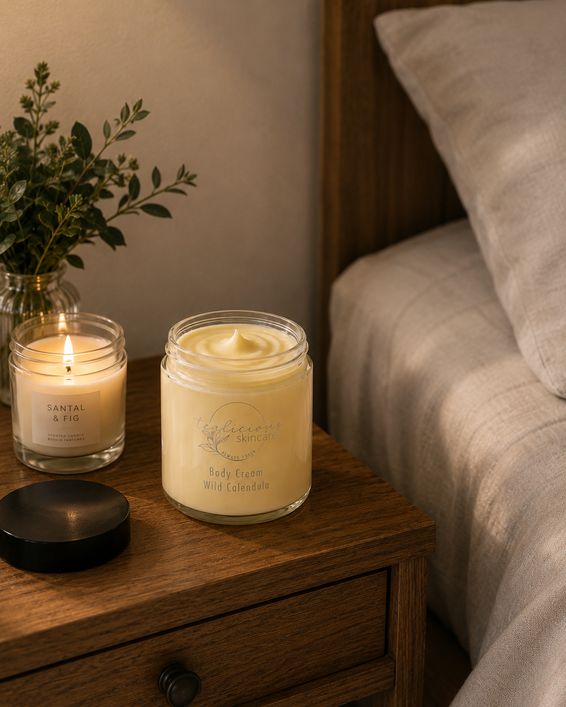 An open glass jar of Tea-licious Skincare Wild Calendula body cream sits on a wooden nightstand next to a lit Santal & Fig candle. This Professional Homestyle shot features soft morning light and realistic shadows, highlighting a relaxing bedside routine for skin barrier support.
