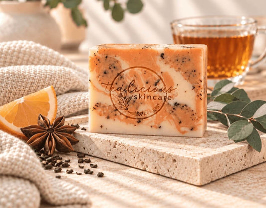 Tea-Licious Spicy Orange Tea Exfoliating Soap Bar with orange and cream swirls and tea flecks, displayed on a stone tray beside orange, star anise and a cup of tea.