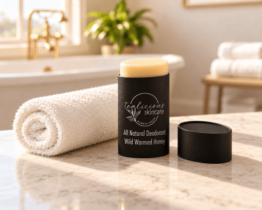 Tea-licious Wild Warmed Honey deodorant in a black oval container with silver lettering, beside its lid and a rolled ivory towel on a sunlit marble bathroom counter.