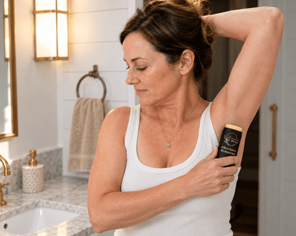 Woman in an ivory sleeveless top applying Tea-licious Wild Warmed Honey deodorant from a black oval container to her underarm in a warmly lit bathroom.