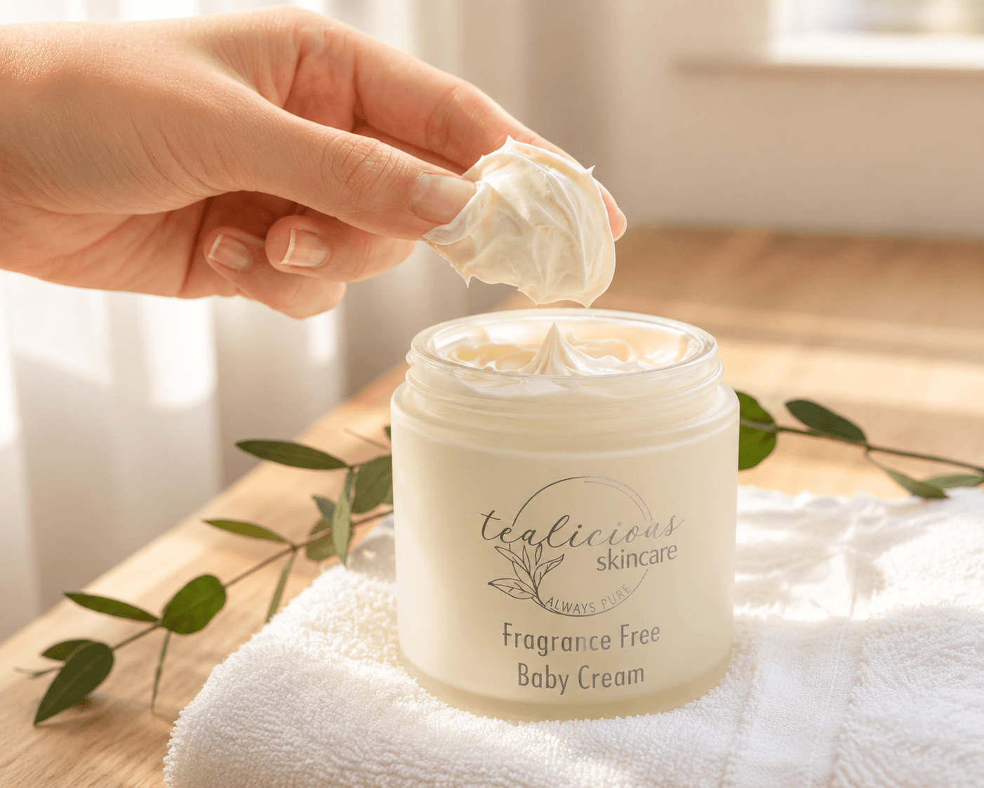 Tea-licious skincare Hand applying cream from a jar labeled 'tealicious skincare' on a wooden surface with a towel and leaves.