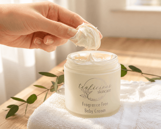Tea-licious skincare Hand applying cream from a jar labeled 'tealicious skincare' on a wooden surface with a towel and leaves.